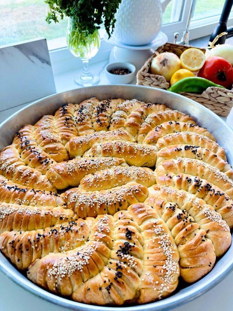 Mediterranean Meat & Cheese Savory Rolls served in a round metal sheet pan that has a spiral pattern with lemon, green pepper, onions, and tomato in a woven basket, a white small bowl with roasted pine nuts, and a glass with fresh herbs for decor on a white countertop.