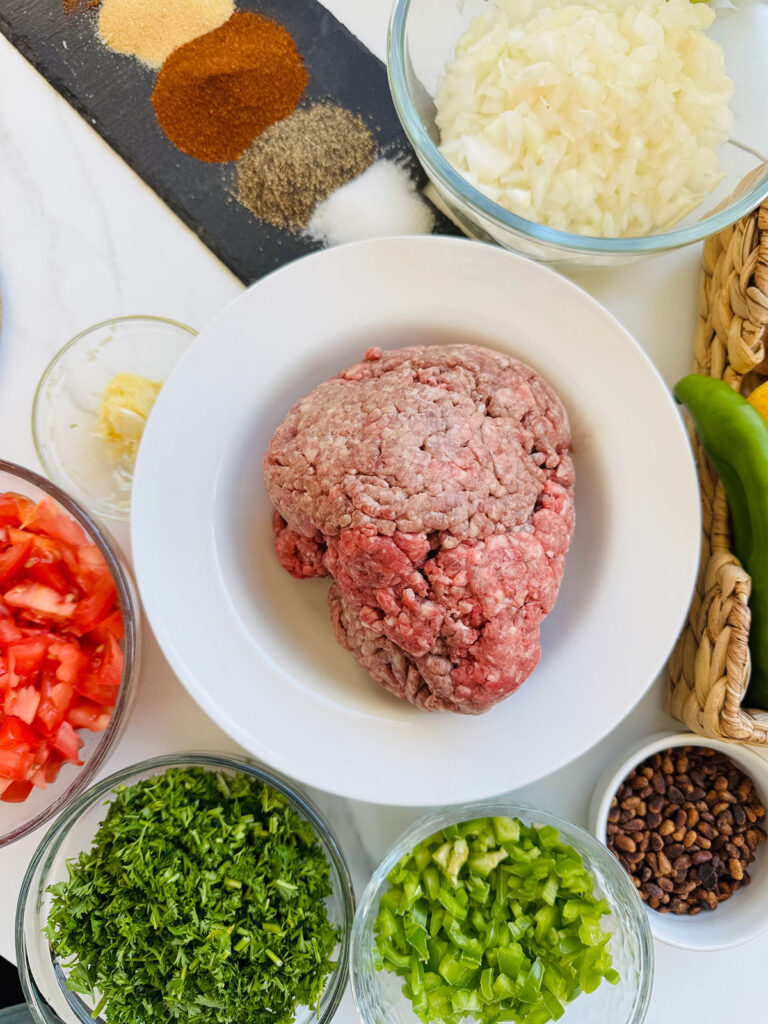 Mediterranean Meat & Cheese Savory Rolls - white bowl with ground beef next to glass bows filled with cheese, chopped tomatoes, cilantro green onions and roasted pine nuts along with salt oregno, paprika and garlic powder on a iron rectangular skillet