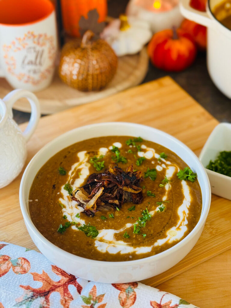 The Best Lentil Soup Recipe - lentil soup in a white bowl on a wooden chopping board with white yogurt drizzled over soup with green chopped fresh parsley as garnish. White pitcher, decorative pumpkins and a fall mug on a round wooden tray in background. Fall towel throwen on wooden board and white container with fresh chopped parsley. 