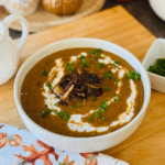 The Best Lentil Soup Recipe - lentil soup in a white bowl on a wooden chopping board with white yogurt drizzled over soup with green chopped fresh parsley as garnish. White pitcher, decorative pumpkins and a fall mug on a round wooden tray in background. Fall towel throwen on wooden board and white container with fresh chopped parsley.
