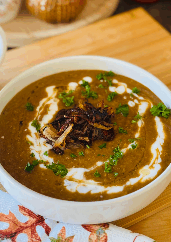 The Best Lentil Soup Recipe - lentil soup in a white bowl on a wooden chopping board with white yogurt drizzled over soup with green chopped fresh parsley as garnish. White pitcher, decorative pumpkins and a fall mug on a round wooden tray in background. Fall towel throwen on wooden board and white container with fresh chopped parsley.