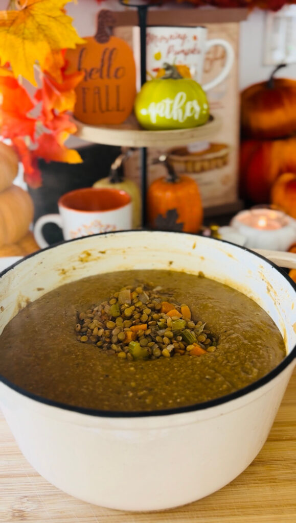 The Best Lentil Soup Recipe - white cast-iron pot with lenitls and veggies in broth on top of a wooden board with a lit candle with decorative pumpkins in background.
