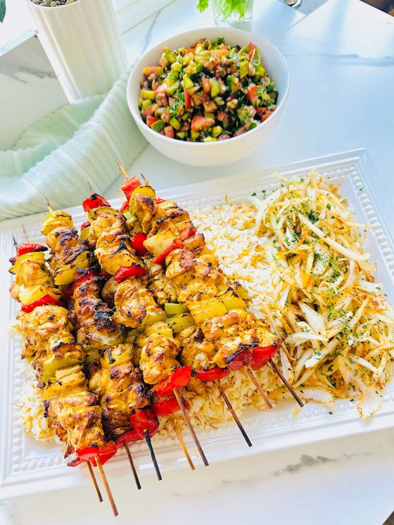 Tender Oven Baked Chicken Kebobs - chicken kebobs ontop of a large white serving platter with rice and seasoned onions with fresh greens sprinkled ontop with a bowl of salad and grey towel in background on a white countertop.