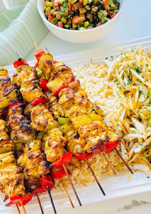 Tender Oven Baked Chicken Kebobs - chicken kebobs ontop of a large white serving platter with rice and seasoned onions with fresh greens sprinkled ontop with a bowl of salad and grey towel in background on a white countertop.