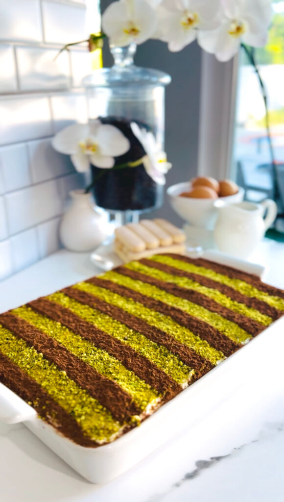 Pistachio Tiramisu Cake – This rich and creamy dessert is made with sweetened pistachio cream and fluffy mascarpone cream, layered to perfection. Finished with a delicate dusting of cocoa powder and crushed pistachios on top. Placed into a white baking platter sitting on a tabletop with ladyfingers in background, a white vase with white a single white orchid strand, a bowl with eggs, white pitcher with a white tiled background.