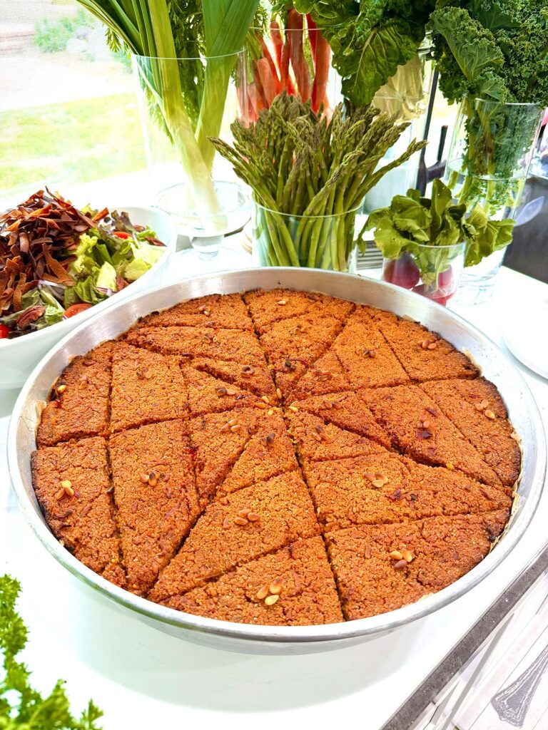 Crispy Lebanese vegan kibbeh baked in a round pan with pine nuts, served with a fresh salad and surrounded by organic vegetables like asparagus, leeks, radishes, carrots, and kale.