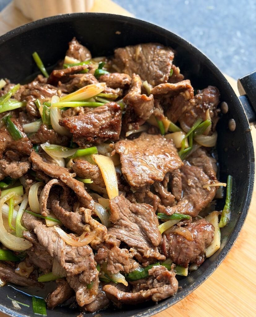 Easy Mongolian Beef - And It's Ready in 30 Minutes! Black wok filled with stir-fried beef, sauted onions, sliced bambo shoots and green onions on top of a wooden chopping board on a black countertop.