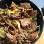 Easy Mongolian Beef - And It's Ready in 30 Minutes! Black wok filled with stir-fried beef, sauted onions, sliced bambo shoots and green onions on top of a wooden chopping board on a black countertop.