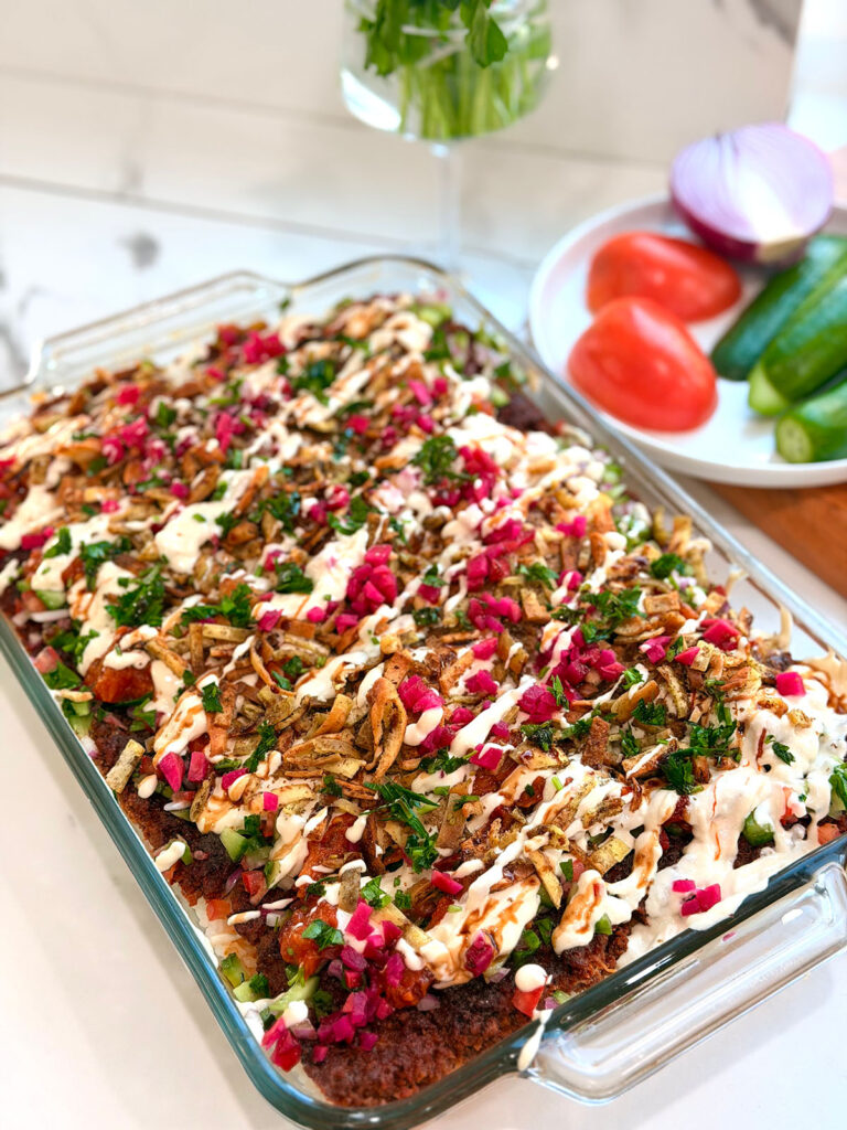 Kebab Crunch Bake - in a glass baking platter on a white countertop with a white plate upper right corner with tomatoes, cucumber slices and a red onion as decor.