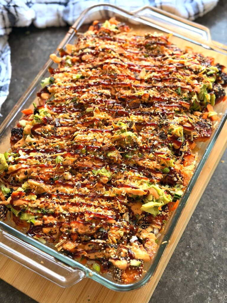 Salmon Baked Sushi Tray- glass platter filled with sushi drizzled with siracha mayo and unagi sushi sauce on a wooden chopping board with a towel at the left corner on a black countertop.