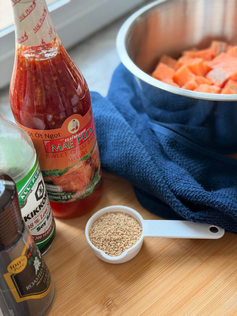 Salmon Baked Sushi Tray - Mae Ploy Sweety Chili Sauce,  Kikkoman soy sauce, sesame oil with a white measuring cup filled with sesame seeds with a blue towel and a metal bowl filled with cubbed salmon chunks on a wooden chopping board.